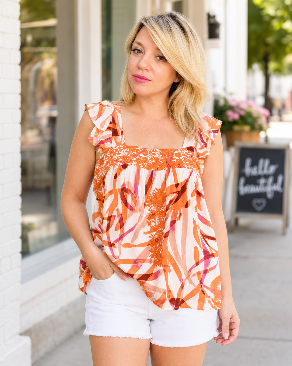 A woman wearing a sleeveless orange and white abstract print top with ruffled shoulders and white shorts, standing on a sidewalk in front of a storefront.