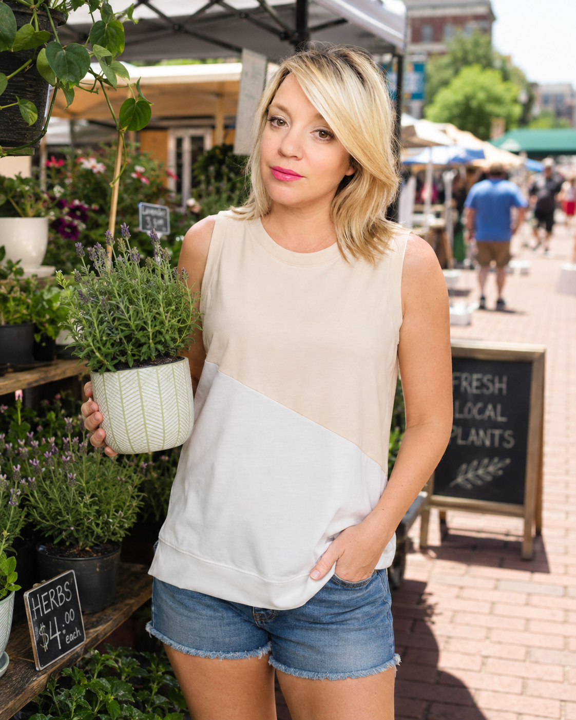 A woman modeling a sleeveless color-block top with a beige upper section and a white lower section, standing at an outdoor farmers market next to a potted plant.