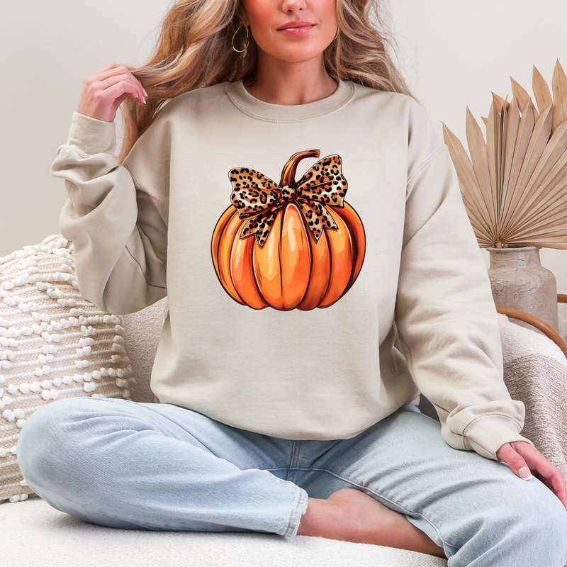 Person wearing a beige sweatshirt with a pumpkin design featuring a leopard print bow, sitting on a white surface.