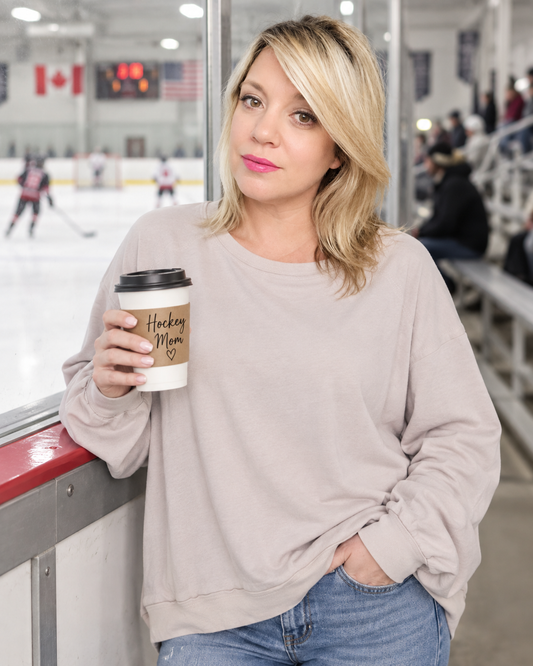 A woman in a light beige crewneck sweatshirt and blue jeans stands at an ice rink barrier, holding a coffee cup with a 'Hockey Mom' design.