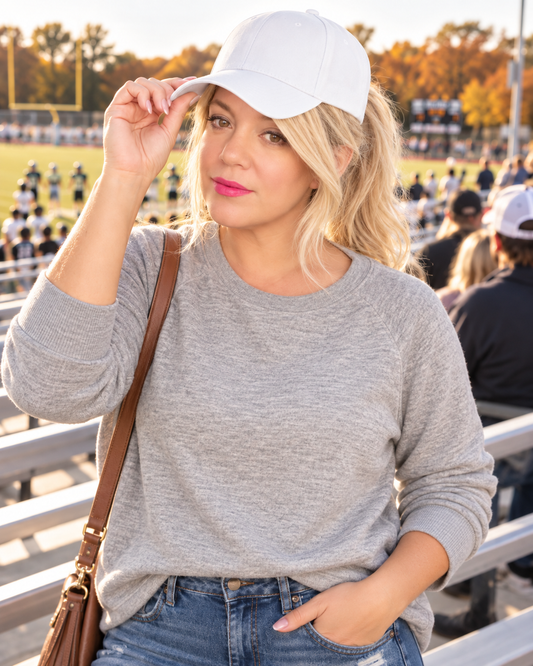A woman in a gray long-sleeve crewneck sweater and white baseball cap stands in a sports stadium, holding her cap with one hand and wearing a brown shoulder bag.