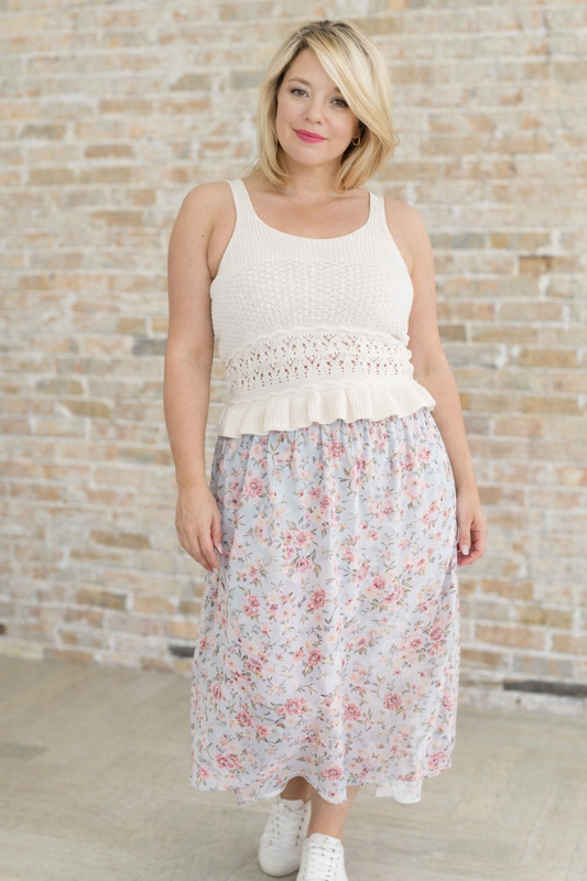 Woman wearing a white sleeveless top with lace details and a floral skirt against a brick wall.