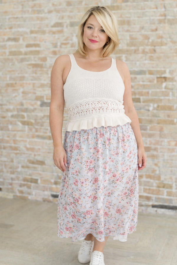 Woman wearing a white sleeveless top with lace details and a floral skirt against a brick wall.